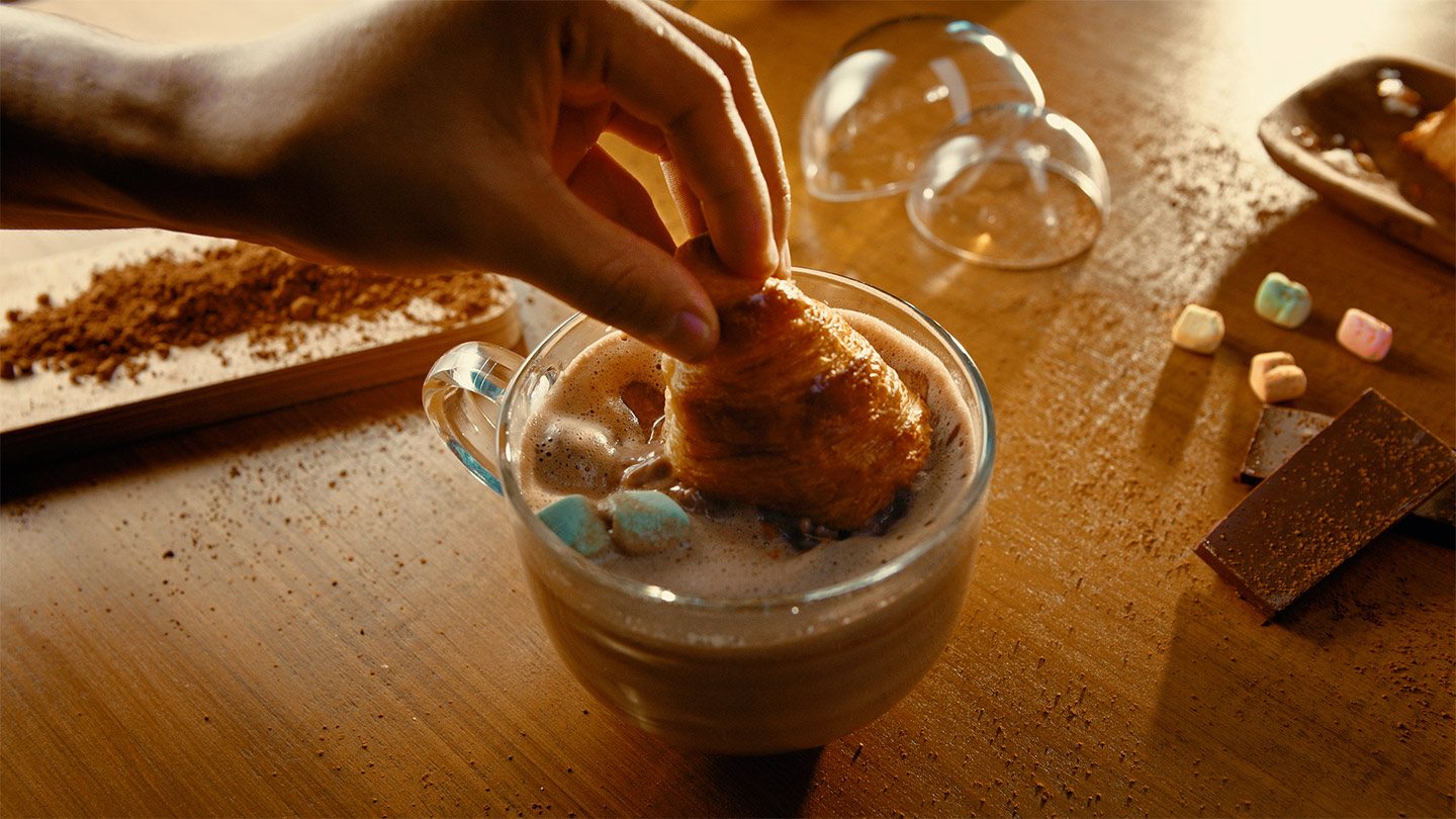 Mano sumergiendo un croissant en una taza de chocolate caliente con malvaviscos en la parte superior.