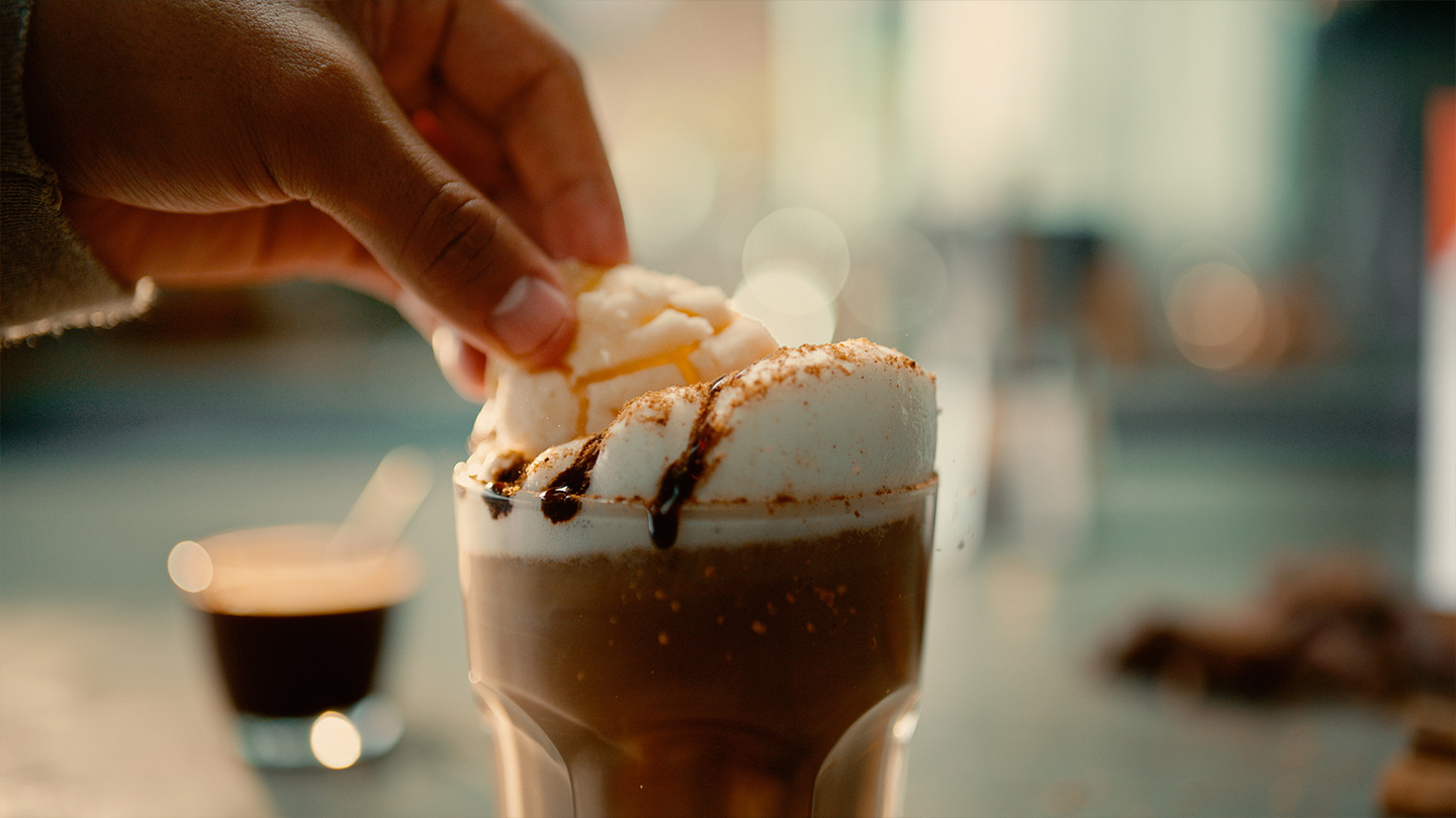 Mano colocando un topping sobre una bebida con abundante espuma y decoración de jarabe de chocolate.