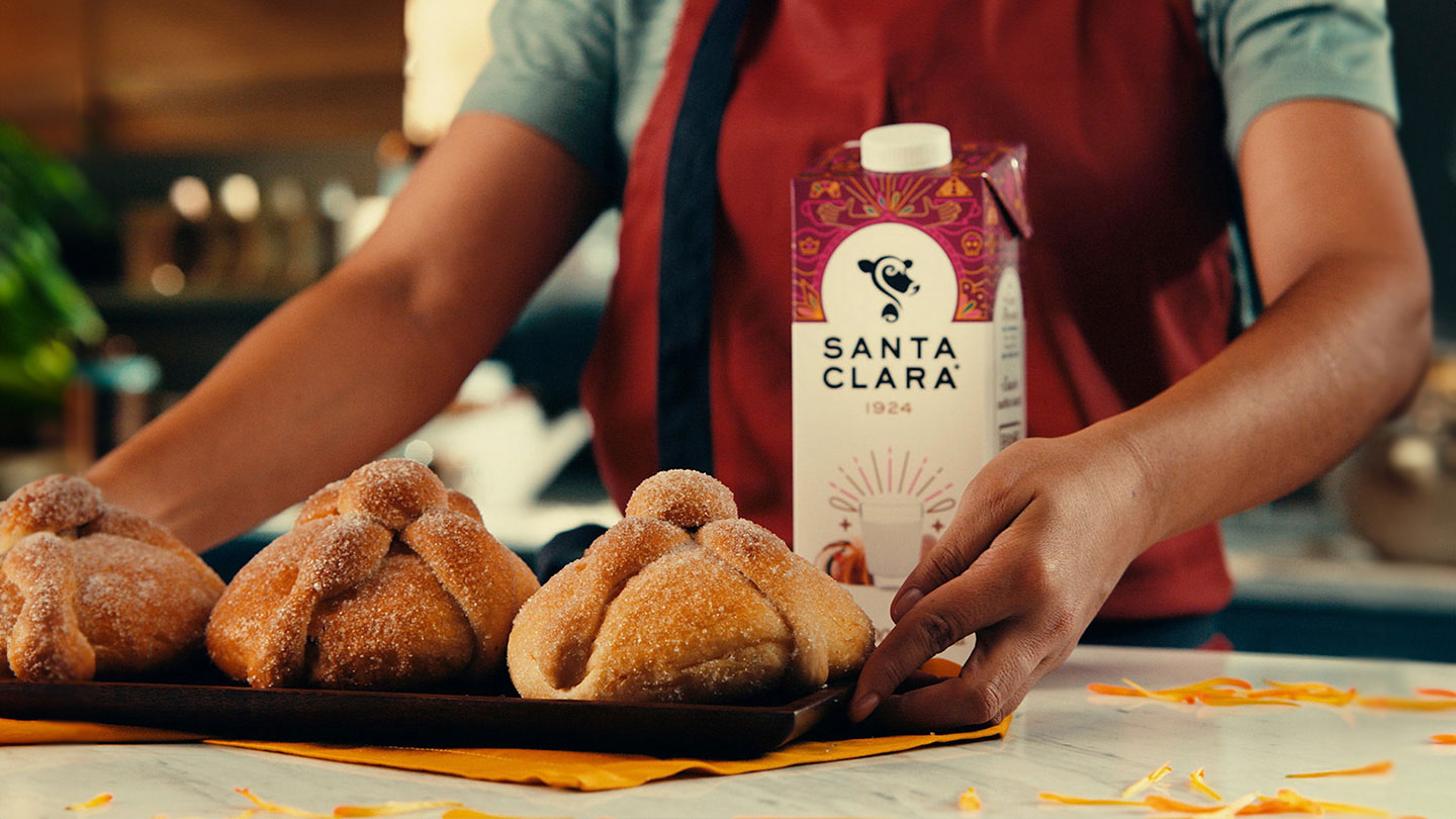 Persona colocando una bandeja con panes de muerto espolvoreados con azúcar en una superficie de mármol, al fondo un cartón de leche Santa Clara con diseño colorido.