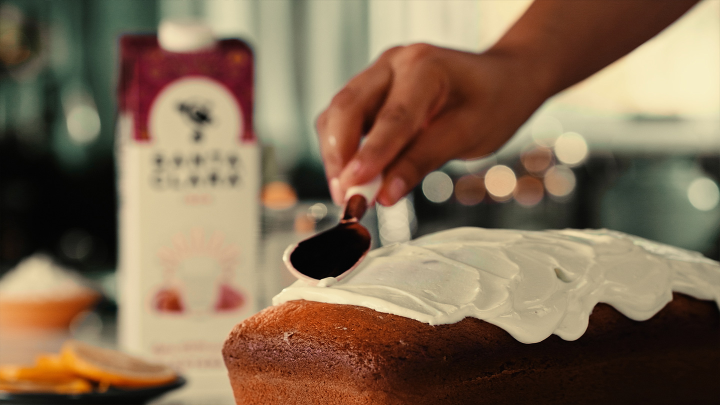 Persona extendiendo glaseado blanco sobre un pastel con una cuchara, con un cartón de leche Santa Clara en el fondo.