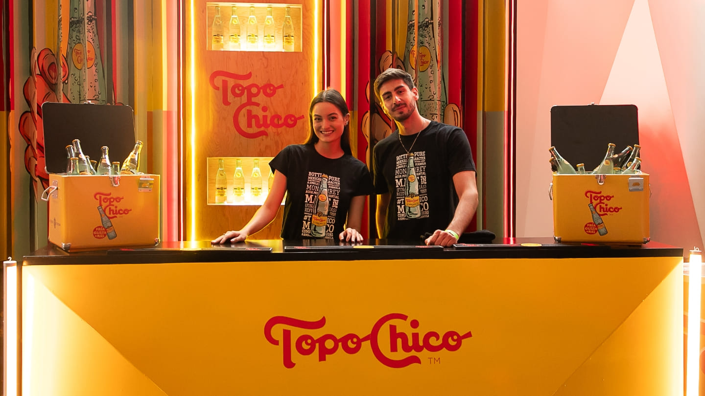 Dos jóvenes detrás del stand de Topo Chico miran a cámara.