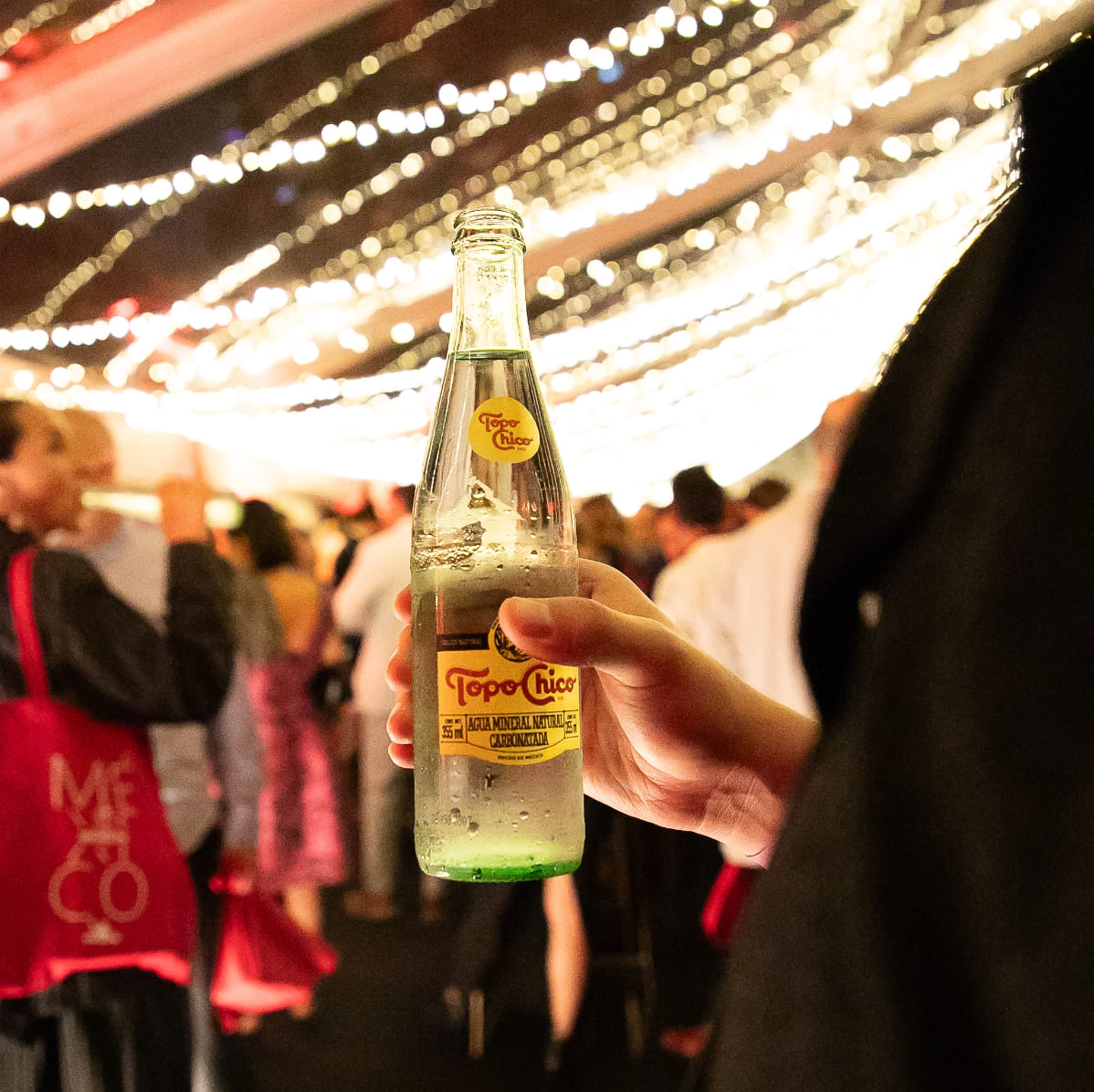 Una mano sostiene una botella de Topo Chico. De fondo se observan personas y luces colgantes. 