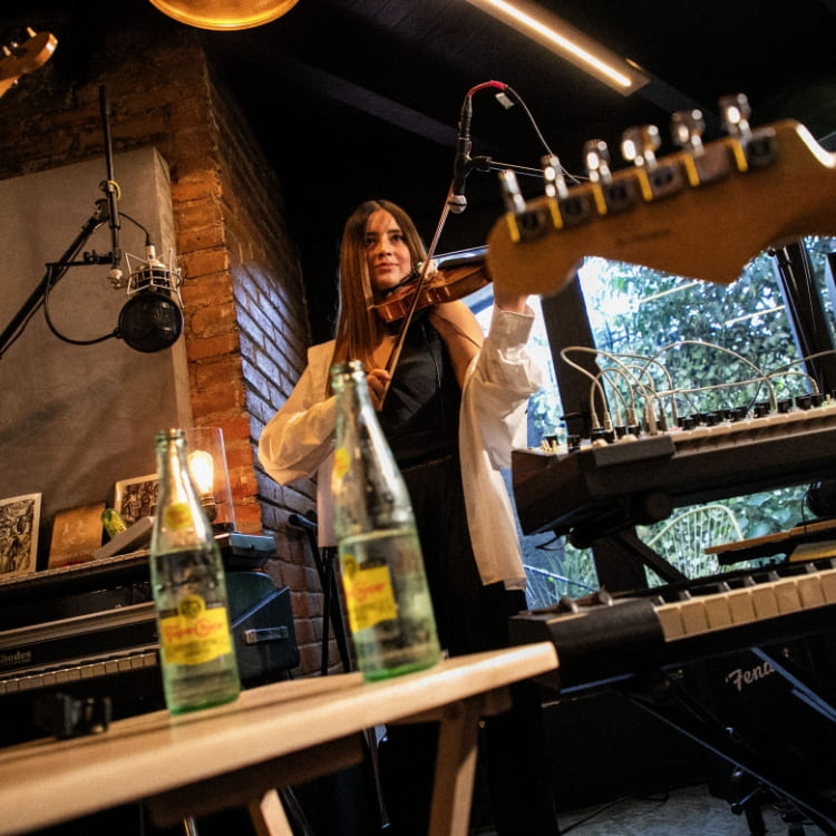 Una joven toca el violín. Delante de ella, se observan dos botellas de Topo Chico sobre una mesa.