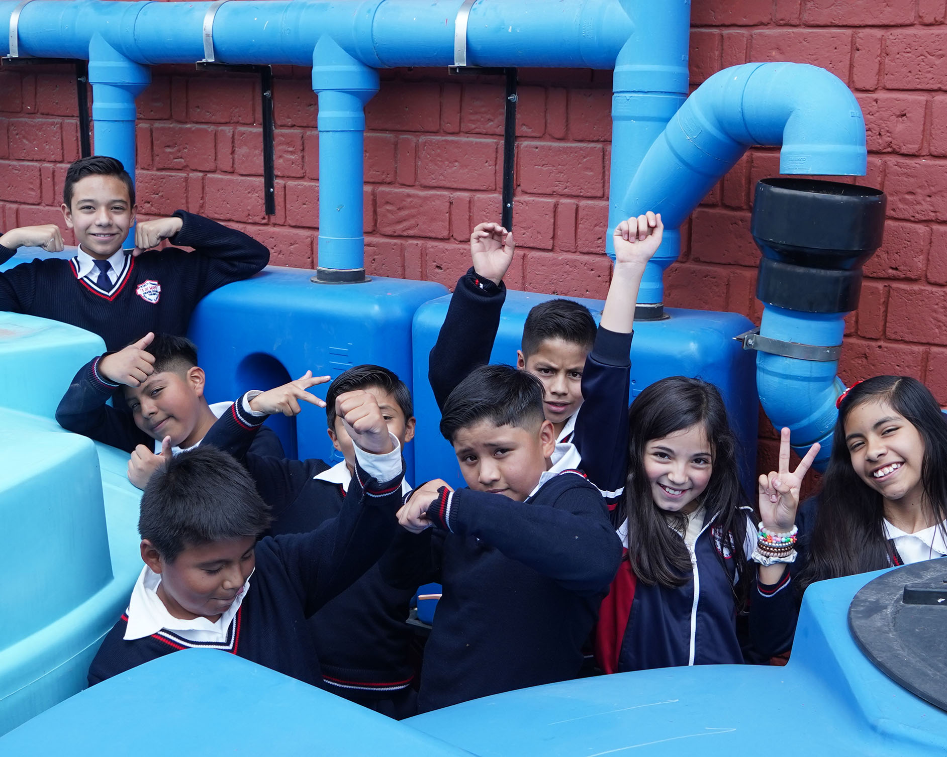 Un grupo de estudiantes en uniformes posando alegremente alrededor de grandes tuberías azules contra una pared de ladrillos rojos. Los niños hacen diversos gestos con las manos y sonríen, creando una escena animada y enérgica.