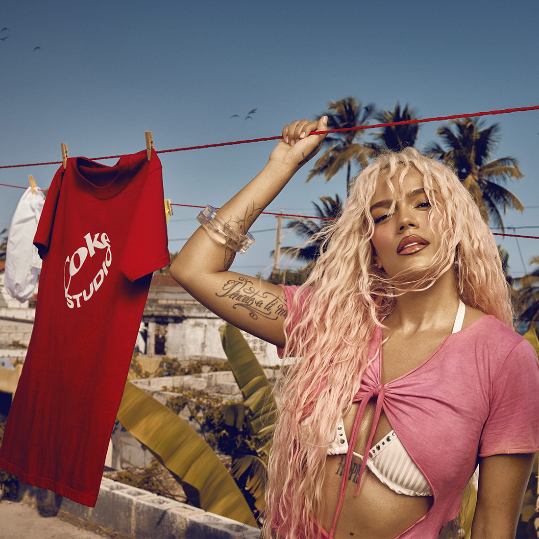 Karol G con el pelo largo y rubio de pie al aire libre cerca de un tendedero con una camiseta roja de Coke Studio colgada. Lleva una parte superior rosa sobre un bikini blanco, mostrando un tatuaje en su brazo. El fondo presenta palmeras y un cielo azul claro