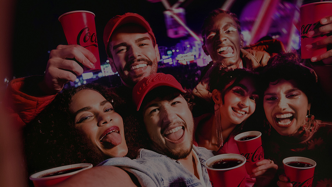 Grupo de jóvenes sonriendo y levantando vasos rojos de Coca-Cola en un ambiente festivo por la noche.