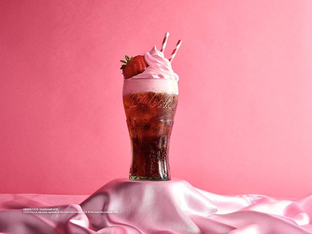 Bebida especial de Coca-Cola con crema batida y decoraciones de colores, presentada en vaso de vidrio sobre base decorativa