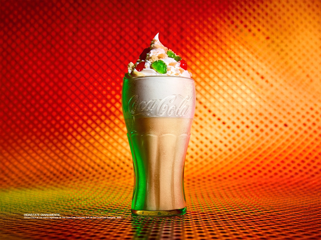 Bebida especial de Coca-Cola con crema batida y decoraciones de colores, presentada en vaso de vidrio sobre base decorativa