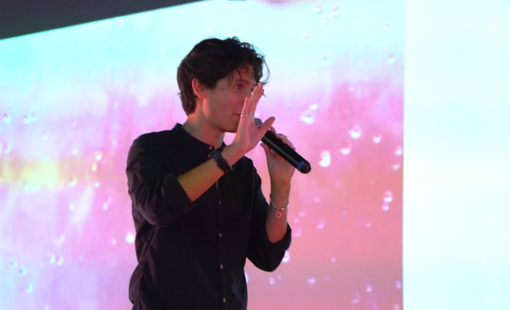 Alexis Omman vestido con camisa negra, hablando o cantando con un micrófono en la mano frente a una pantalla de fondo colorida con tonos rosados y amarillos.