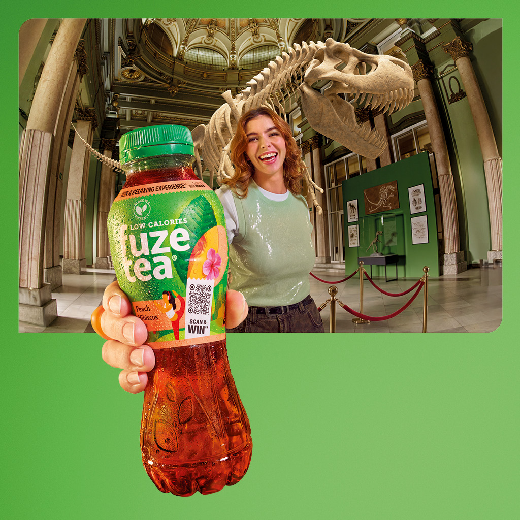 Persoon houdt een fles Fuze Tea vast in een museum met een dinosaurusskelet op de achtergrond. 
