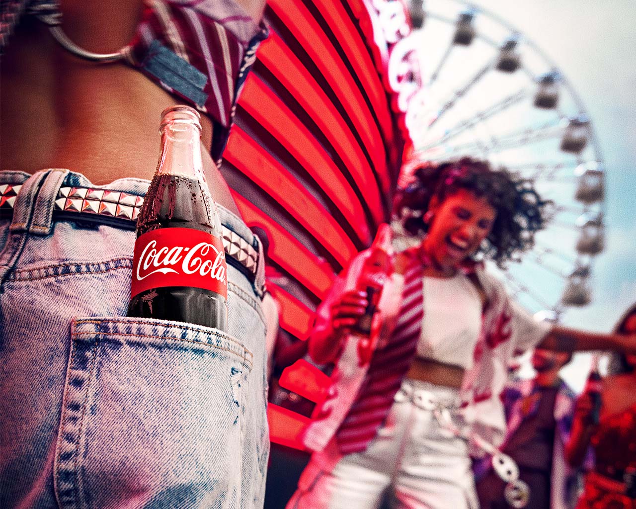 Un primer plano de una botella de Coca-Cola asomando del bolsillo trasero de unos Jeans desgastados, con el fondo de una feria iluminada y personas disfrutando del ambiente festivo.