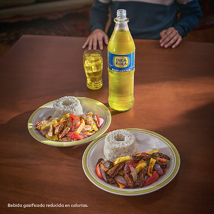 Sobre una mesa de madera, están dispuestos dos platos de comida. Uno con Saltado de Pollo, y otro con Saltado de Carne. Están acompañados por una botella retornable de Inca Kola y un vaso de vidrio con Inca Kola en su interior.