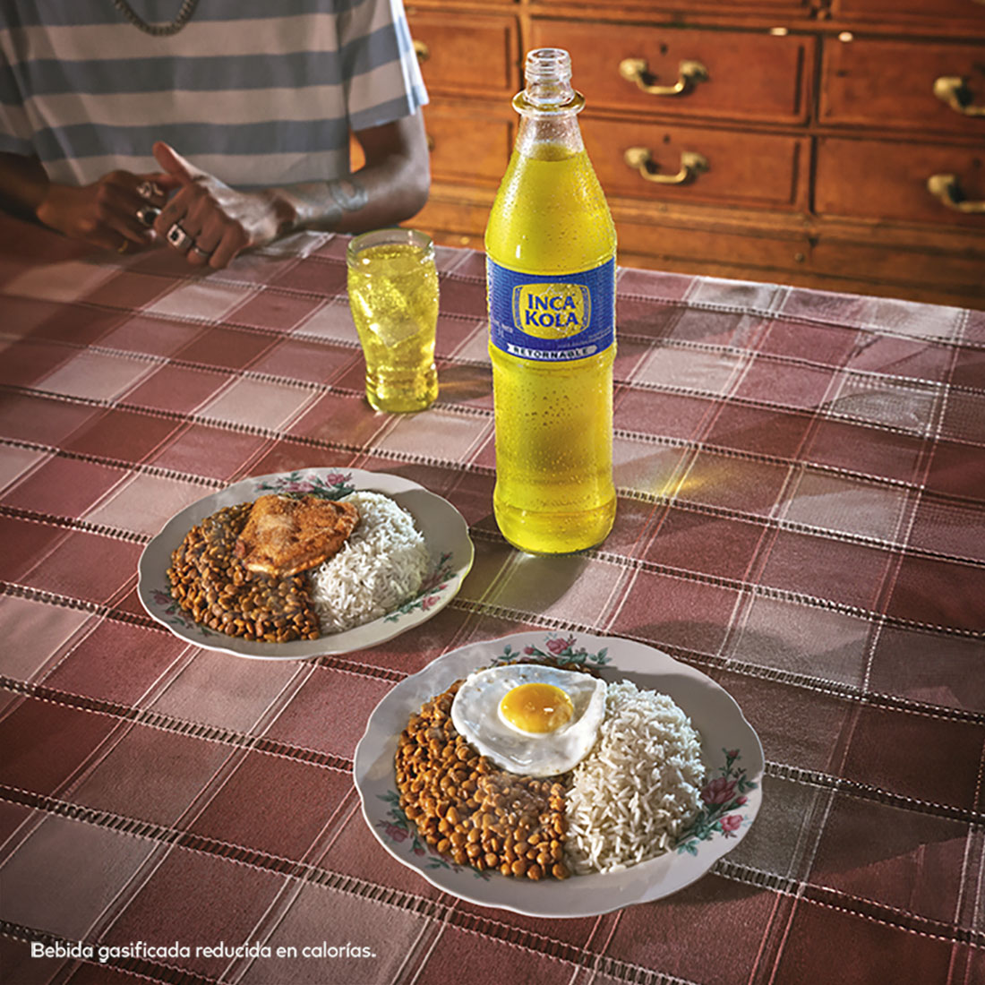 Sobre una mesa están dispuestos dos platos de comida. Uno con Lentejas con Milanesa, y otro con Lentejas con Huevo. Están acompañados por una botella retornable de Inca Kola, y un vaso de vidrio con Inca Kola en su interior.