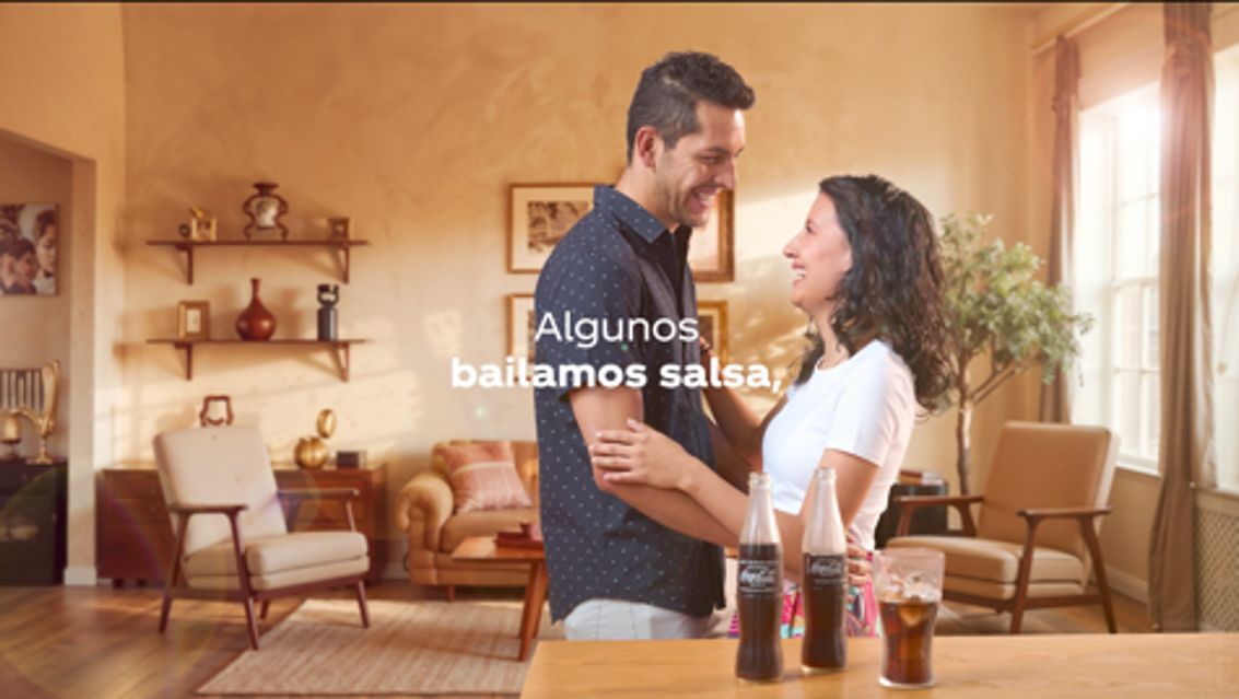 Pareja sonriendo y bailando en una sala de estar con dos botellas de Coca-Cola sobre la mesa. Sobre la imagen, el título  "Algunos bailamos salsa"