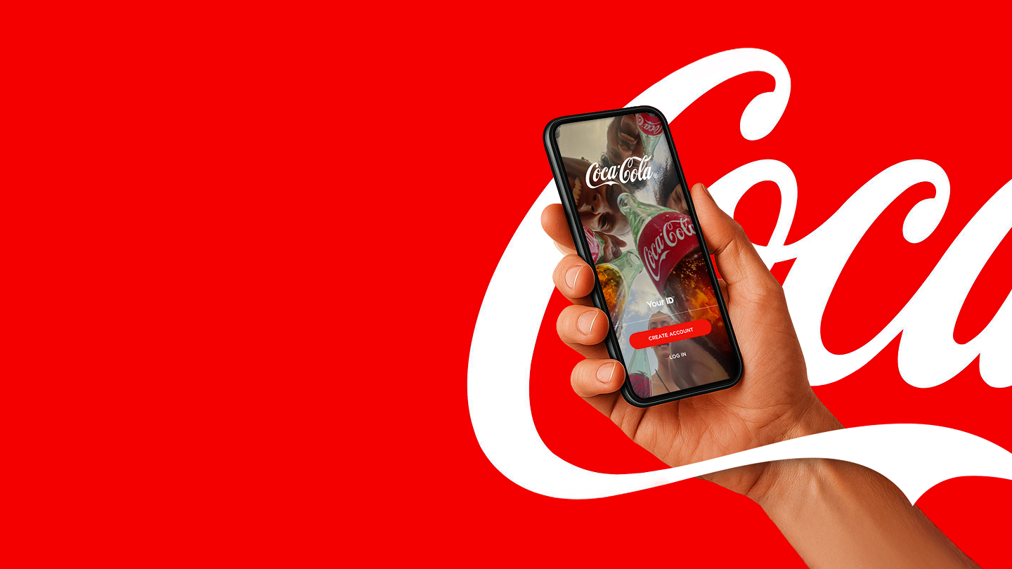 Osoba sedi za stolom sa tastaturom i monitorom, drži mobilni telefon i igra igricu, pored teksta koji poziva na preuzimanje Coca-Cola aplikacije sa ponudama, nagradama, igrama, lojalti programima i vestima.