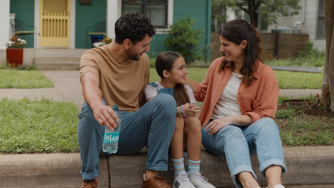 A mother and father sit with their child on a curb in their neighborhood, smiling.