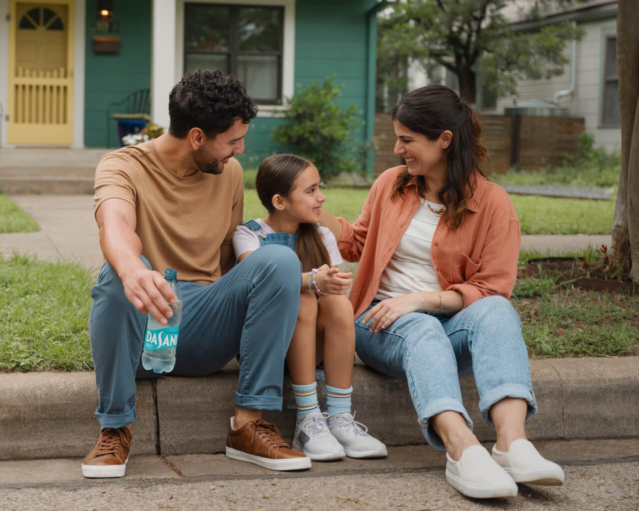 A mother and father sit with their child on a curb in their neighborhood, smiling.