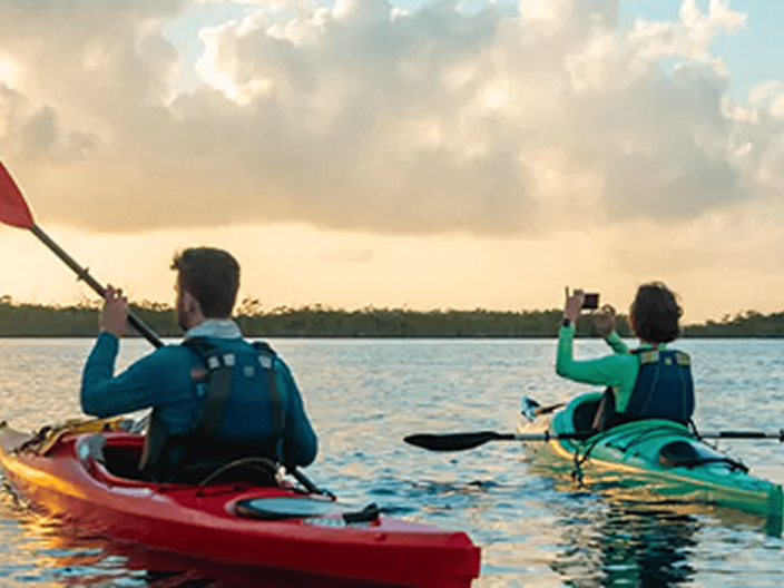 Two people kayaking on calm water at sunset, one in a red kayak paddling and the other in a green kayak taking a photo