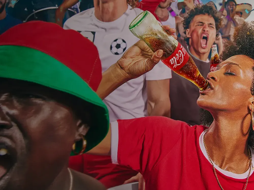 A female soccer fan in a red shirt drinks from a glass Coca-Cola bottle while surrounded by a cheering crowd.