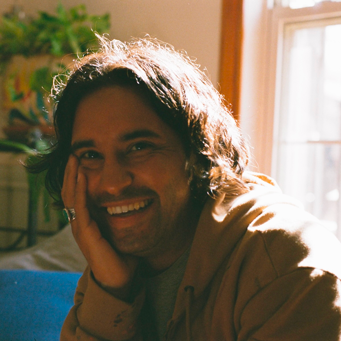 A warm, sunlit profile photo of the smiling male mural artist, resting his head on his hand in an indoor setting.