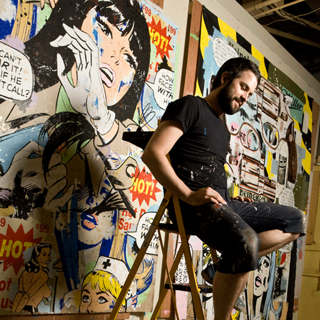 Pop‑art mural artist seated on ladder in painted clothes inside studio filled with large comic‑style wall art panels.
