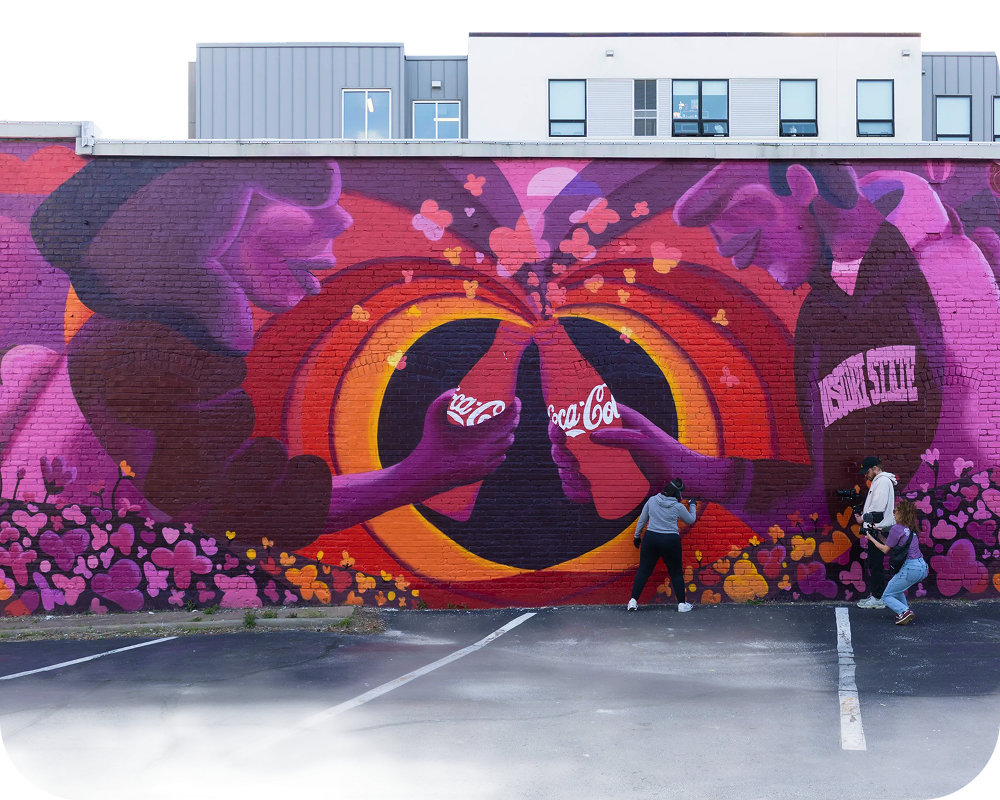 Street art scene with team painting the Paint the Nation Coca-Cola mural across a parking lot wall filled with hearts and color