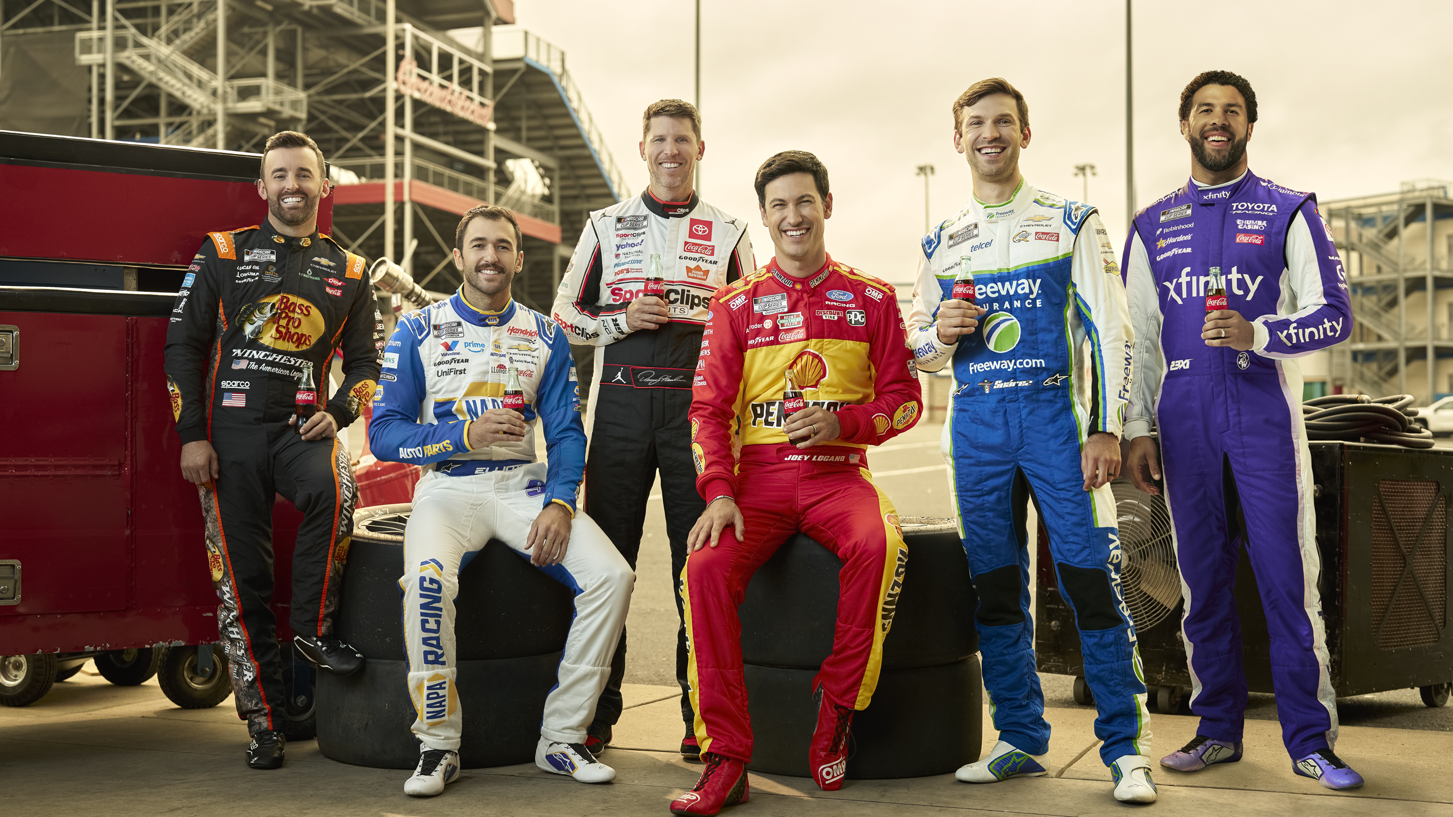 Group of NASCAR drivers in colorful race suits holding Coca‑Cola bottles at the track, highlighting Coca‑Cola NASCAR 500 race day experience.
