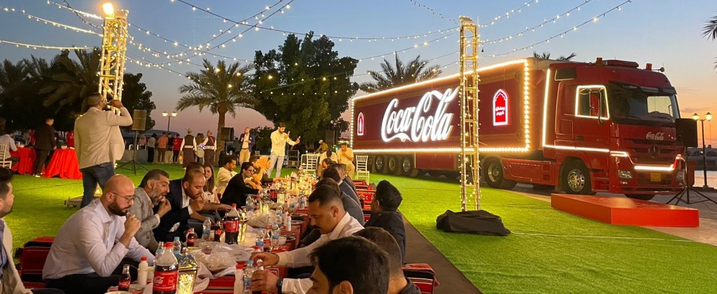 A large group of individuals eating on a long table in the open air beside a Coca-Cola truck with themed lightning